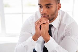 Lost in thoughts. Thoughtful young African man in shirt and tie holding hands on chin and looking away while sitting at his working place photo