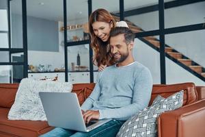 Cheerful young couple using laptop and smiling while spending time at home photo