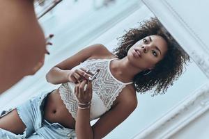 maquillaje perfecto. reflejo de una atractiva joven africana sosteniendo un pintalabios mientras está de pie frente al espejo en casa foto