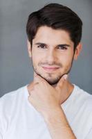 Ready to melt your heart. Confident young man holding hand on chin and looking at camera while standing against grey background photo