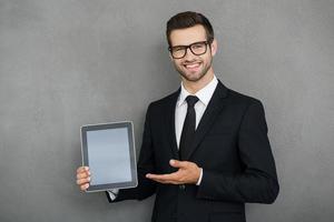 Copy space on his tablet. Cheerful young businessman holding digital tablet and pointing at it while standing against grey background photo