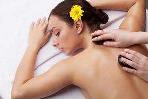 Stone treatment. Top view of attractive young woman with flower in head lying on front and keeping eyes closed while massage therapist massaging her back with spa stones photo