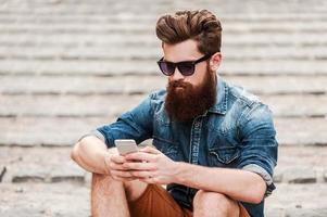 mantenerse conectado. guapo joven barbudo hombre sujetando el teléfono móvil mientras está sentado al aire libre foto