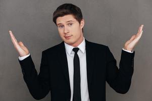 Feeling uncertain. Frustrated young man in formalwear stretching out hands and looking at camera while standing against grey background photo