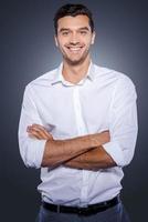 Confident and successful. Happy young man in white shirt looking at camera and keeping arms crossed while standing against grey background photo