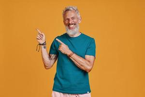 Happy senior man pointing copy space and smiling while standing against orange background photo