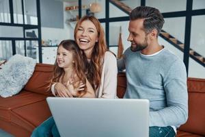 Playful beautiful family with little girl bonding together and smiling while using laptop at home photo