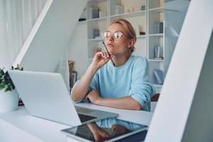 Beautiful young woman using laptop and looking thoughtful while working in office photo