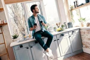 Thinking about something. Handsome young man in casual wear drinking wine and using his smart phone while sitting in the kitchen at home photo