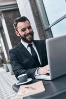 Achieving great results. Good looking young man in full suit using computer and smiling while sitting in the cafe outdoors photo