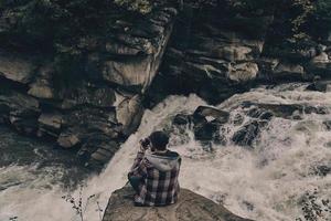 Risky job. Top view of young modern man photographing while sitting on the rock with the river below photo