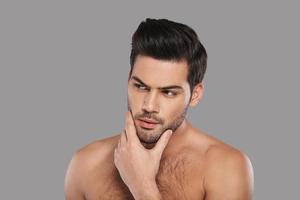 What if... Thoughtful young man looking away and keeping hand on chin while standing against grey background photo