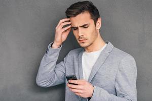 Everyday issues. Pensive young man holding mobile phone and looking at it while standing against grey background photo