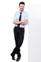 Leaning at the copy space. Full length of handsome young man in shirt and tie leaning at the copy space and keeping arms crossed while standing against white background photo