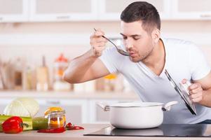 sopa de degustación. apuesto joven probando sopa de la sartén mientras está de pie en la cocina foto