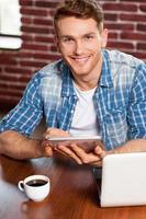 Examining his new tablet. Top view of handsome young man working on digital tablet and smiling while sitting at the table photo