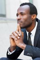 Lost in thoughts. Thoughtful young African man in formalwear keeping hands clasped and looking away while sitting outdoors photo