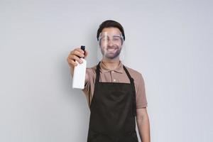 hombre joven guapo en delantal con protector facial y sosteniendo spray de limpieza mientras está de pie contra el fondo gris foto