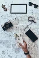 Preparing to travel. Close up top view of man holding passport and money with smart phone, sunglasses, photo camera, compass, magnifying glass lying on map around