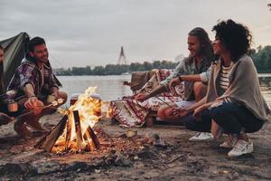 No cares. Group of young people in casual wear roasting marshmallows over a campfire while resting near the lake photo