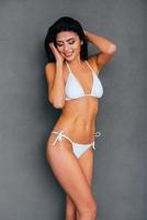 confiada en su cuerpo perfecto. Atractiva mujer sonriente joven en bikini blanco posando sobre fondo gris foto