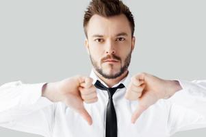 My decision. Portrait of confident young man in shirt and tie looking at camera and showing his thumbs down while standing against grey background photo