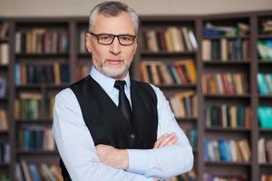 Confident and intelligence. Confident grey hair senior man in formalwear keeping arms crossed and looking at camera while standing against bookshelf photo