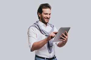 Using modern technologies. Beautiful young man in smart casual clothes working on digital tablet while standing against grey background photo