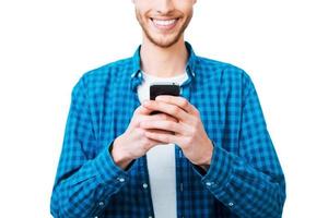 Choosing right words. Close-up of young man in shirt holding mobile phone and smiling while standing against white background photo