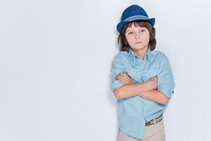 lleno de inspiración. niño pequeño con sombrero y manteniendo los brazos cruzados mientras está de pie contra el fondo blanco foto