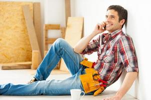 Handyman relaxing. Handsome young handyman talking on the mobile phone and smiling while sitting on the floor photo