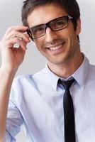 Expert look. Portrait of handsome young man in shirt and tie adjusting his eyeglasses and smiling while standing against grey background photo