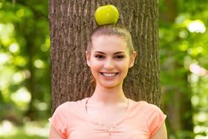 en armonía con la naturaleza. bella joven que lleva manzana en la cabeza y mira la cámara con una sonrisa mientras se apoya en el árbol en un parque foto