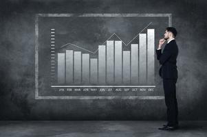 Thinking about solution. Full length of thoughtful young man in full suit standing near concrete wall and looking at the illustrated chart photo