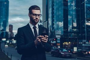 Quick business message. Night time image of confident young businessman in formalwear holding smart phone and looking at it while standing outdoors with cityscape in the background photo