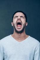 Man shouting. Furious young man keeping eyes closed and mouth open while standing against grey background photo