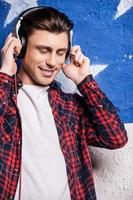 Listening to his favorite music. Cheerful young man in checked shirt keeping eyes closed and adjusting his headphones  while standing against background photo