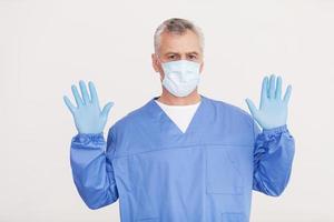 cirujano confiado. médico senior de pelo gris con mascarilla quirúrgica mirando a la cámara y mostrando sus manos con guantes azules mientras está aislado en blanco foto