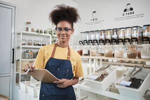 Portrait of young Black female shopkeeper smiling and looking at camera arranging natural products at refill store, zero-waste grocery, and plastic-free, environment-friendly with reusable containers. photo