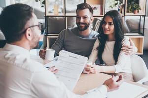 You just need to sign it. Happy young couple sitting together at the desk and looking at man sitting in front of them and holding contract photo