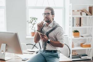 On the way to success. Handsome young man holding note pad and looking thoughtful while sitting on the desk at his working place in creative office photo