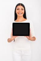 Copy space on her tablet. Cheerful young woman stretching out digital tablet and smiling while standing against white background photo