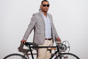 mi bicicleta es mi estilo de vida. un joven africano seguro de sí mismo con gafas de sol apoyado en su bicicleta de estilo retro y mirando a la cámara mientras se enfrenta a fondo blanco foto