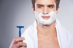 Shaving with bad razor. Young shirtless man looking at the razor and expressing negativity while standing isolated on grey background photo