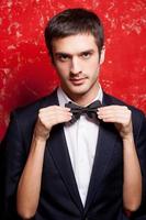 Macho in bow tie. Handsome young man in formalwear standing against red background while woman hiding behind him and adjusting his bow tie photo