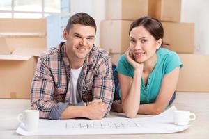 Just moved in. Cheerful young couple lying on the floor with a blueprint on it while cardboard boxes laying on the background photo