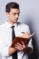 Writing down his ideas. Confident young man in shirt and tie writing something in note pad while standing against grey background photo