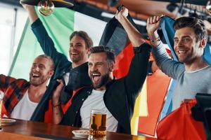 Cheering young men covered in international flags enjoying beer while watching sport game in the pub photo
