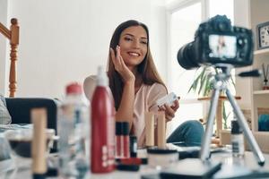 Beautiful young woman applying lip gloss and smiling while making social media video photo