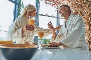 Beautiful mature couple in bathrobes enjoying breakfast together while spending time in the domestic kitchen photo